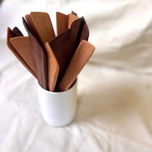 K&ouml;nnte beinhalten: Eine Sammlung von K&uuml;chenutensilien aus Holz in verschiedenen Braunt&ouml;nen, von hellem Beige bis zu dunklem Walnuss. Die Utensilien sind in einem wei&szlig;en Keramikbecher angeordnet. Der Hintergrund ist ein weicher, wei&szlig;er Stoff.