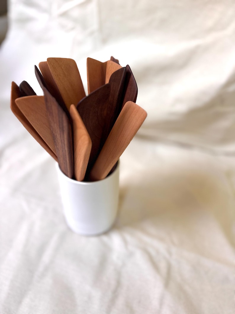K&ouml;nnte beinhalten: Eine Sammlung von K&uuml;chenutensilien aus Holz in verschiedenen Braunt&ouml;nen, von hellem Beige bis zu dunklem Walnuss. Die Utensilien sind in einem wei&szlig;en Keramikbecher angeordnet. Der Hintergrund ist ein weicher, wei&szlig;er Stoff.