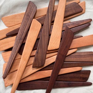 K&ouml;nnte beinhalten: Eine Sammlung von Holzbuttermessern in verschiedenen Braun- und Beiget&ouml;nen. Die Messer haben einen langen, spitzen Griff und eine breite, flache Klinge. Die Holzmaserung ist sichtbar. Die Messer sind in einem Haufen angeordnet.