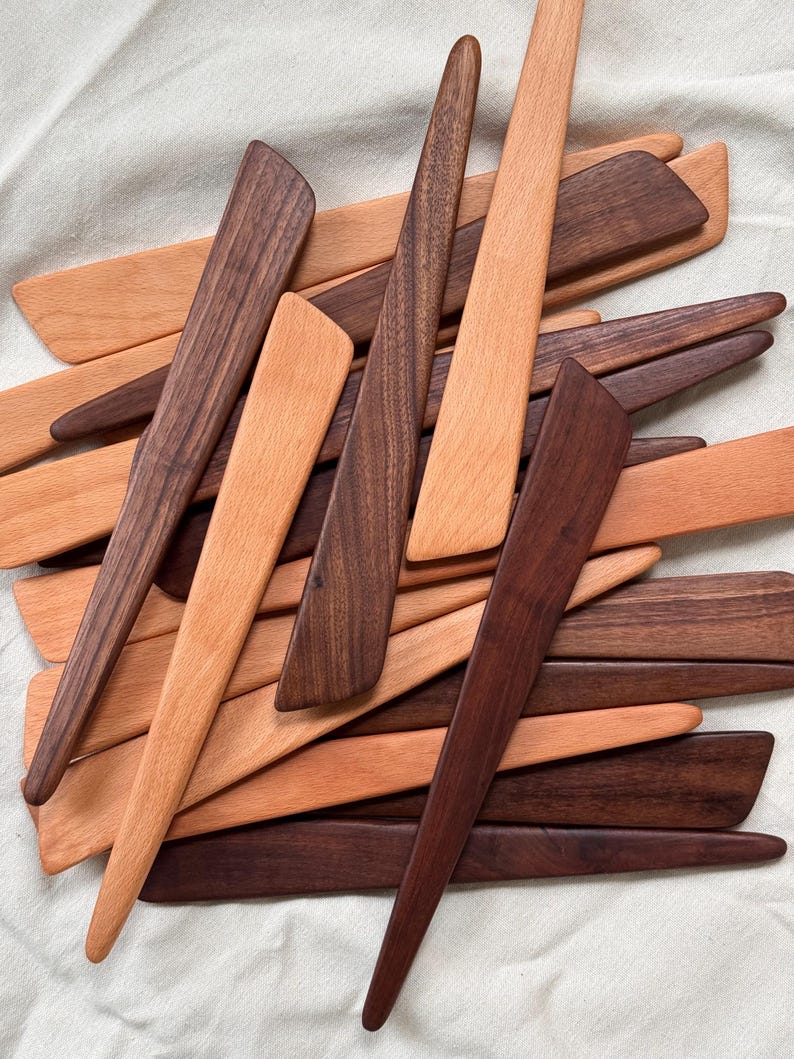K&ouml;nnte beinhalten: Eine Sammlung von Holzbuttermessern in verschiedenen Braun- und Beiget&ouml;nen. Die Messer haben einen langen, spitzen Griff und eine breite, flache Klinge. Die Holzmaserung ist sichtbar. Die Messer sind in einem Haufen angeordnet.