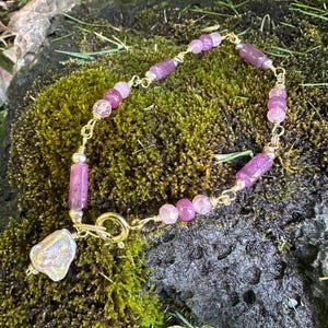 May include: A gold-toned wire-wrapped bracelet featuring a series of faceted pink and purple beads, cylindrical purple stones, and small silver-toned spacers. The bracelet includes a circular clasp and a single irregular white baroque pearl charm.