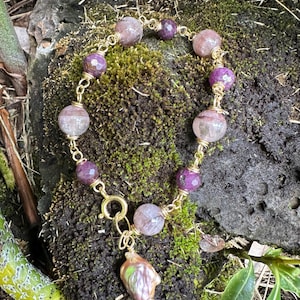 May include: A gold-toned chain bracelet featuring alternating faceted purple and translucent pinkish-grey beads, connected by gold wire loops. A single irregular, iridescent, light-colored pendant hangs from a circular gold clasp at the bottom.