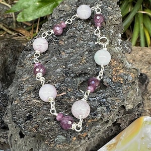May include: A bracelet featuring alternating round light lavender beads and smaller faceted deep purple beads connected by silver-toned metal links. The jewelry piece includes a circular clasp mechanism and is arranged in a circular shape.
