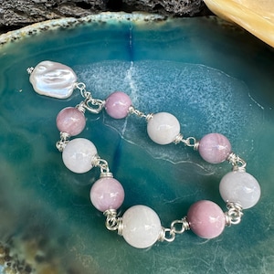 May include: A beaded bracelet featuring alternating round mauve and translucent white stone beads connected by silver-toned wire links, finished with a single irregular white baroque pearl charm attached to a silver-toned metal clasp.