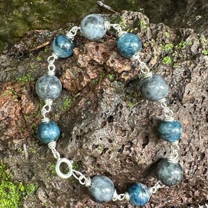 Puede incluir: Una pulsera con cuentas redondas alternas en tonos gris oscuro y azul profundo, conectadas por eslabones de alambre plateado. Las cuentas tienen un acabado pulido y liso, dispuestas en un patrón circular con un pequeño cierre circular plateado.