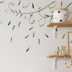 May include: A white shelf unit with three shelves, each containing various items. The top shelf has a white jar with black polka dots, a wooden bunny, and a woven basket. The middle shelf has a wooden sign with the text "Your Design Here", a wooden block with the number 20, and a string of wooden beads. The bottom shelf has a white ceramic pitcher, a woven basket, a white fabric box with a black panda face, and a woven basket. The wall behind the shelves has a green vine decal with leaves.