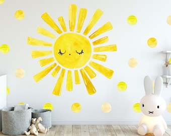 May include: A yellow watercolor sun with a smiling face and closed eyes, surrounded by yellow and cream watercolor dots on a white wall. The sun is a wall decal and the dots are wall stickers.