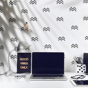 May include: A white desk with a laptop, a notebook that says "GOOD VIBES ONLY." and a lamp. The wall behind the desk has a repeating pattern of gray chevron shapes.