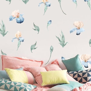 May include: A white bed with a pink blanket and several pillows. The pillows are in various colors and patterns, including yellow, blue, and green. The wall behind the bed is decorated with watercolor-style iris flower decals in shades of blue and pink.