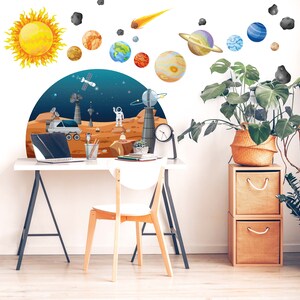 May include: A white desk with a black metal frame and a white chair. The desk has a laptop computer, a potted plant, and a clock on it. The wall behind the desk has a colorful solar system wall decal with planets, stars, and a comet.