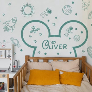 May include: A white wall with a teal Mickey Mouse head silhouette with the name "Oliver" inside. The wall is decorated with space-themed stickers, including a rocket, planets, stars, and astronauts.