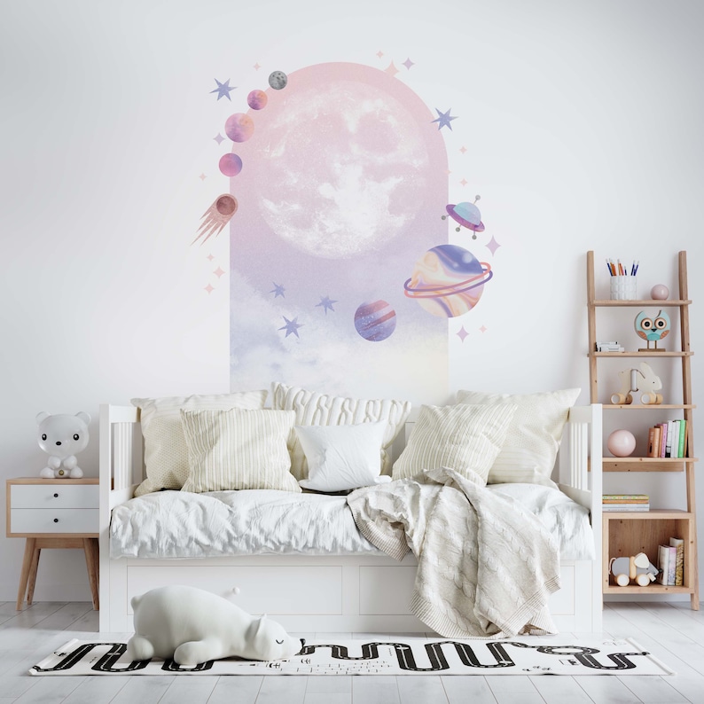 May include: A white daybed with a white and gray blanket and pillows. The wall behind the bed has a large pink and purple moon and stars decal. There is a small wooden shelf with books and toys to the right of the bed.