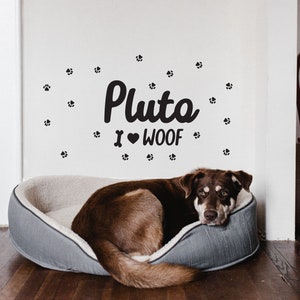 May include: A brown dog is lying in a grey dog bed. The dog is looking at the camera. The wall behind the dog has a black paw print decal and the text "Pluto I ♥ WOOF".