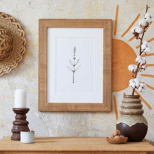 Puede incluir: Un dibujo en blanco y negro de una planta enmarcado en un marco de madera marr&oacute;n. El dibujo est&aacute; colgado en una pared con un sombrero tejido, una vela blanca, un candelero marr&oacute;n, un candelero de cer&aacute;mica blanca, un jarr&oacute;n de cer&aacute;mica marr&oacute;n con un tallo de algod&oacute;n y una cesta tejida.