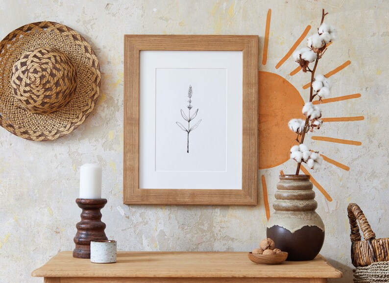 Puede incluir: Un dibujo en blanco y negro de una planta enmarcado en un marco de madera marr&oacute;n. El dibujo est&aacute; colgado en una pared con un sombrero tejido, una vela blanca, un candelero marr&oacute;n, un candelero de cer&aacute;mica blanca, un jarr&oacute;n de cer&aacute;mica marr&oacute;n con un tallo de algod&oacute;n y una cesta tejida.