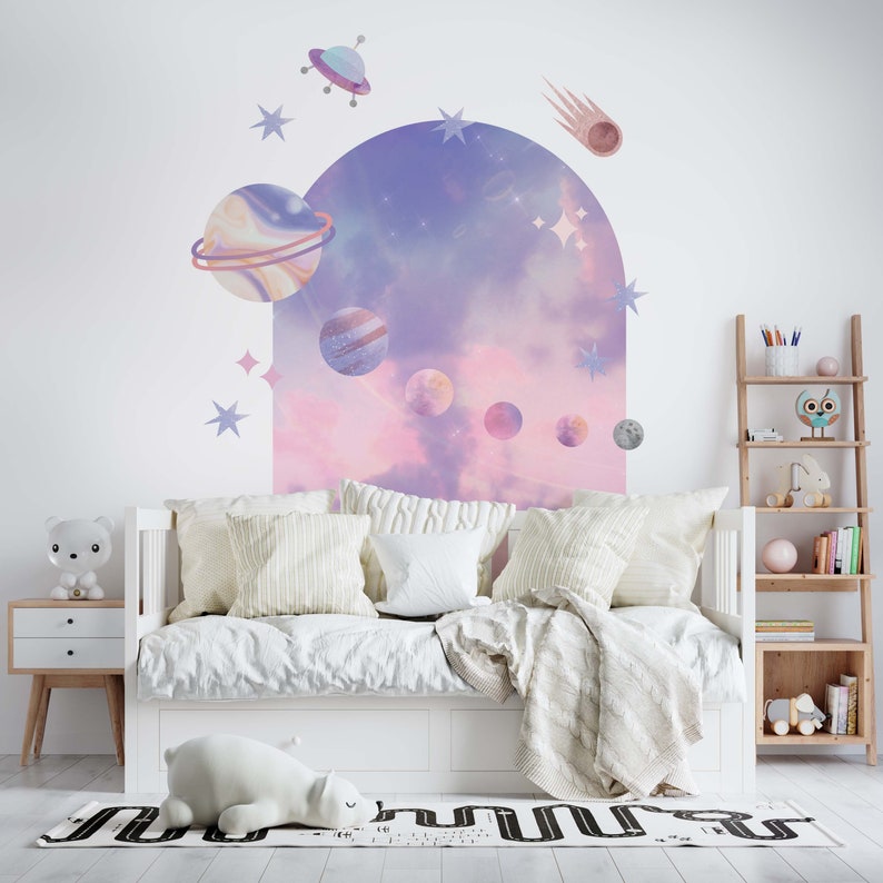 May include: A white bed with a pink and purple galaxy themed wall decal. The decal features planets, stars, a comet, and a spaceship. The bed has a white blanket and pillows.