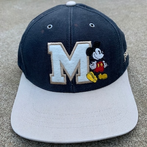 May include: A navy blue and beige baseball cap with a white felt letter "M" and a Mickey Mouse character embroidered on the front.