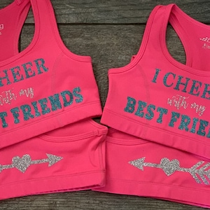 May include: Two pink sports bras with the text "I cheer with my best friends" in teal glitter. Each bra has a silver glitter arrow with a heart in the center.
