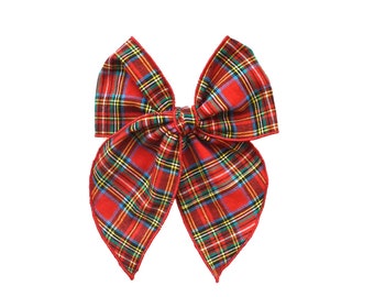 May include: A fabric hair bow featuring a red, green, yellow, and blue plaid tartan pattern. The bow has four distinct, pointed loops gathered at the center with a knotted fabric band, and the edges are finished with a thin red trim.