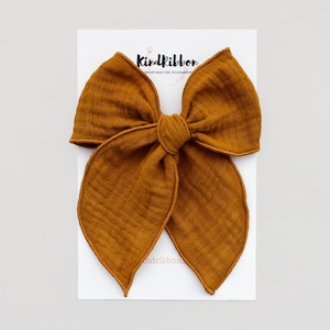 May include: A large mustard yellow fabric bow with a stitched edge. The bow is on a white card with the text "Kindribbon Handmade Hair Accessories".
