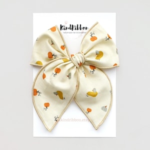 May include: A large cream-colored bow with a pumpkin print. The bow has a brown stitched edge and is tied in a large, full bow.