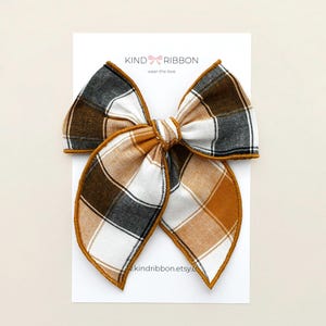 May include: A large plaid hair bow in shades of brown, white, and black, with a gold-colored trim. The bow is centered on a white card with the text "KIND RIBBON" and the website address.