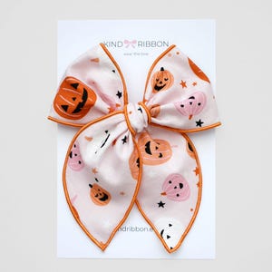 May include: A light pink hair bow featuring a Halloween pumpkin and star design. The bow has orange edging and is knotted in the middle. The bow is on a white card with the text "KIND RIBBON" and "wear the love".