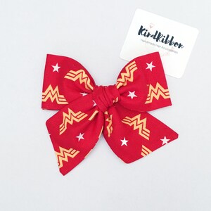 K&ouml;nnte beinhalten: Eine rote Stoffschleife mit einem Wonder Woman Logo-Muster und wei&szlig;en Sternen. Die Schleife ist in der Mitte gebunden und hat auf jeder Seite zwei Schlaufen. Ein wei&szlig;es Etikett mit dem Text "Kindribbon Handmade Hair Accessories" befindet sich im Hintergrund.