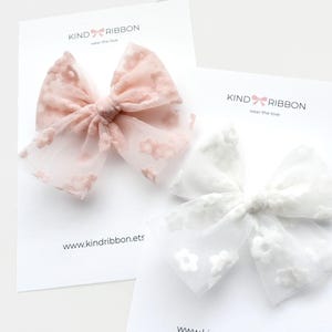 Op de afbeelding: Twee transparante haarstrikken, één in blush roze en één in wit, elk versierd met bloemenborduurwerk. De strikken worden gepresenteerd op witte kaarten met de tekst "KIND RIBBON" en een websiteadres.