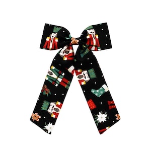 May include: Black Christmas-themed bow with a festive pattern of nutcrackers, gingerbread men, and Christmas stockings. The bow is tied with a small, round, gingerbread man accent in the center. White polka dots are scattered across the black background.