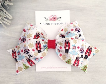 May include: A hair bow made of white ribbon with a repeating pattern of nutcrackers, ballerinas, Christmas trees, gold snowflakes, and red peppermint candies. The bow has a solid red center knot and is attached to a white card labeled KIND RIBBON.