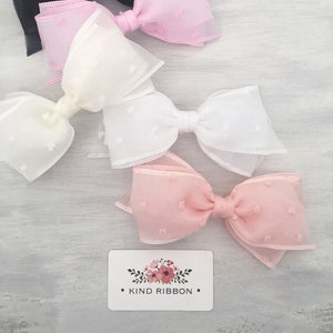 May include: Four hair bows in different colors: black, pink, white, and light pink. The bows are made of sheer fabric with small white dots. The bows are arranged in a row on a white wooden surface. The text "KIND RIBBON" is visible on a white card with a floral design.