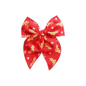 K&ouml;nnte beinhalten: Eine rote Stoffschleife mit einem sich wiederholenden Muster des Wonder Woman-Logos und wei&szlig;en Sternen. Die Schleife ist in der Mitte gebunden und hat zwei Schlaufen und zwei B&auml;nder. Das Wonder Woman-Logo ist goldfarben.