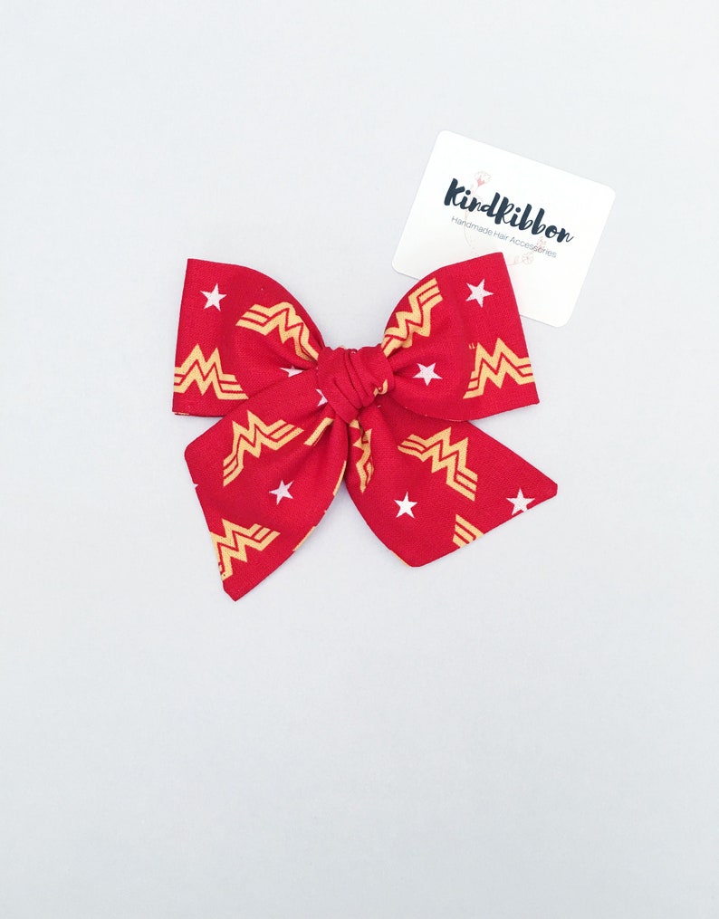 K&ouml;nnte beinhalten: Eine rote Stoffschleife mit einem Wonder Woman Logo-Muster und wei&szlig;en Sternen. Die Schleife ist in der Mitte gebunden und hat auf jeder Seite zwei Schlaufen. Ein wei&szlig;es Etikett mit dem Text "Kindribbon Handmade Hair Accessories" befindet sich im Hintergrund.