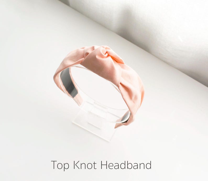 May include: A light pink fabric headband with a knotted top. The headband is on a clear display stand.