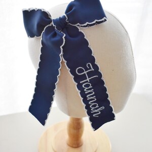 Peut inclure: Un n&oelig;ud pour cheveux bleu marine en ruban gros-grain avec des bords festonn&eacute;s blancs. Le n&oelig;ud porte le pr&eacute;nom Hannah brod&eacute; en &eacute;criture cursive blanche sur l'un des longs pans tombants. Le ruban est nou&eacute; en forme de n&oelig;ud classique.