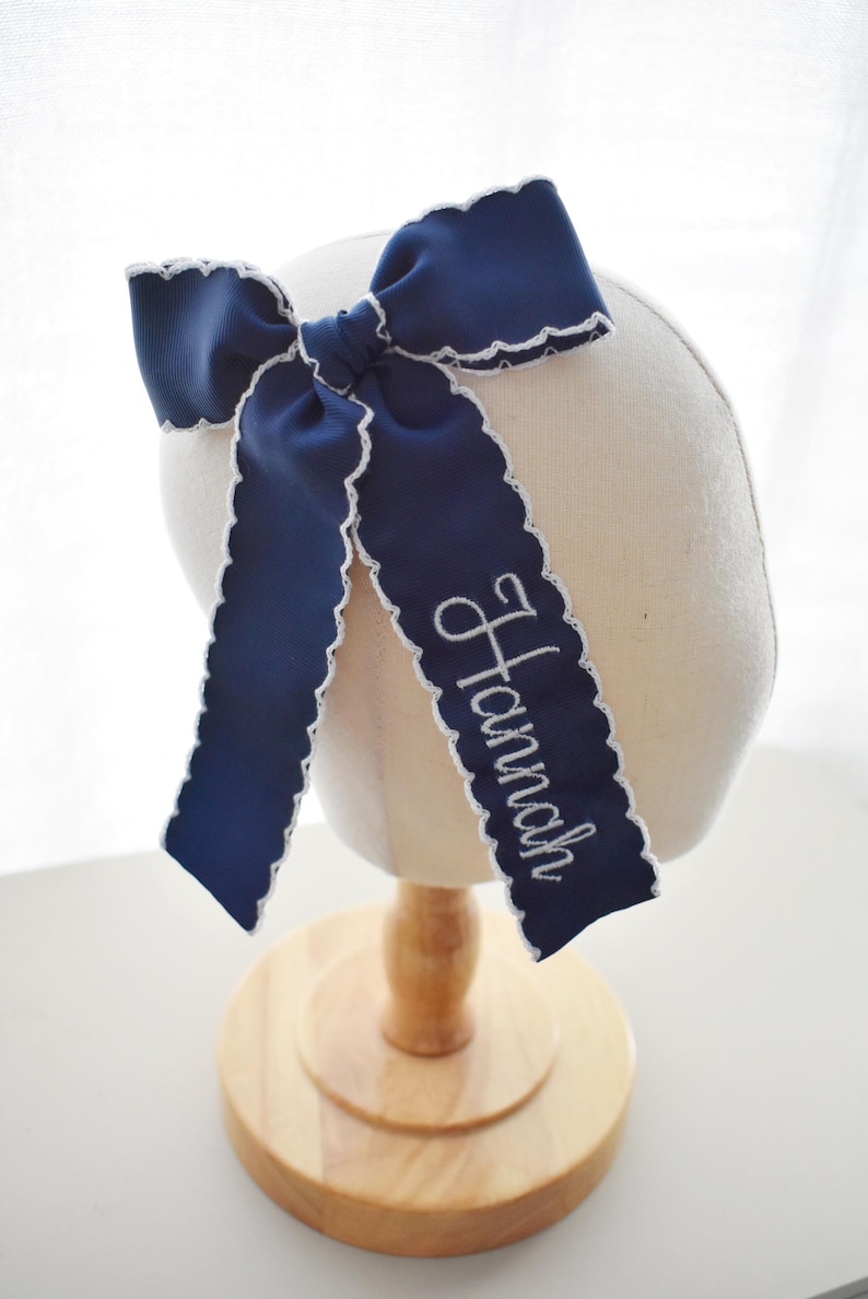 Peut inclure: Un n&oelig;ud pour cheveux bleu marine en ruban gros-grain avec des bords festonn&eacute;s blancs. Le n&oelig;ud porte le pr&eacute;nom Hannah brod&eacute; en &eacute;criture cursive blanche sur l'un des longs pans tombants. Le ruban est nou&eacute; en forme de n&oelig;ud classique.