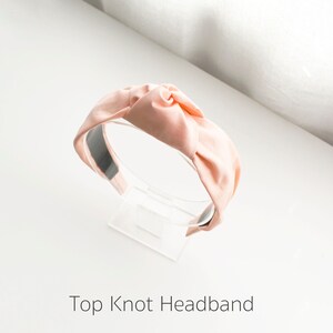 May include: A light pink fabric headband with a large knot on top. The headband is on a clear display stand.