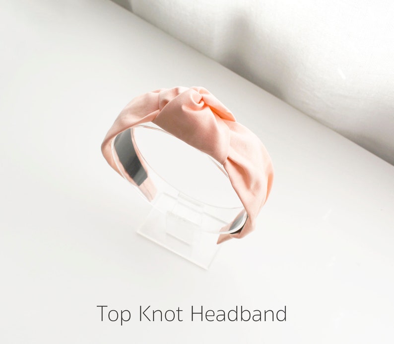 May include: A light pink fabric headband with a large knot on top. The headband is on a clear display stand.