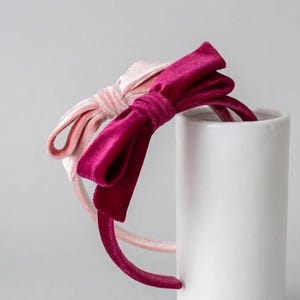 Puede incluir: Dos diademas de terciopelo, una en color magenta intenso y otra en rosa pálido, cada una con un lazo prominente en el lateral. Las diademas están confeccionadas con una tela suave y afelpada y se muestran apiladas una sobre la otra.