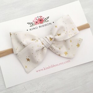 May include: A white fabric headband with a gold star pattern and a large bow. The headband is on a white card with the text "KIND RIBBON" and "www.kindribbon.etsy.com".