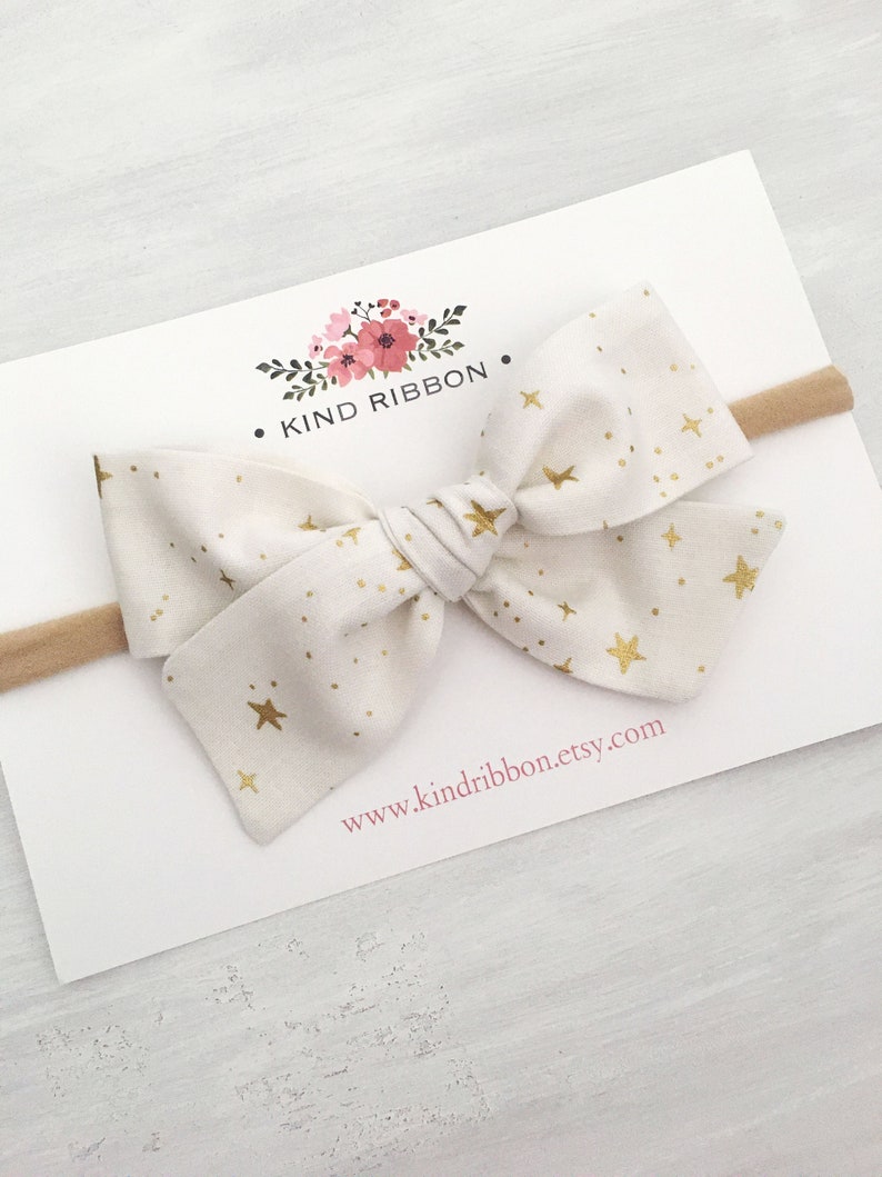 May include: A white fabric headband with a gold star pattern and a large bow. The headband is on a white card with the text "KIND RIBBON" and "www.kindribbon.etsy.com".