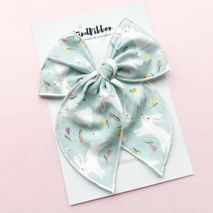 May include: A large, light blue fabric bow with a white bunny and floral pattern. The bow is on a white card with the text "birdribbon Handmade Hair Accessories". The background is a soft pink.