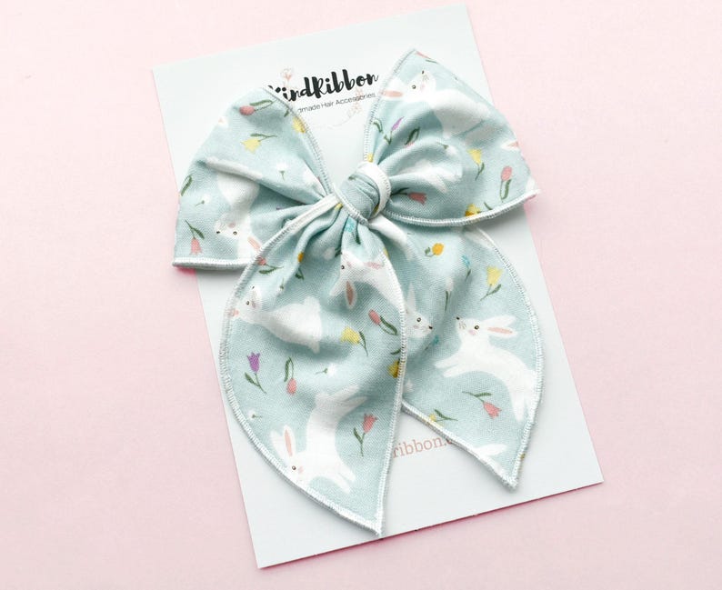 May include: A large, light blue fabric bow with a white bunny and floral pattern. The bow is on a white card with the text "birdribbon Handmade Hair Accessories". The background is a soft pink.