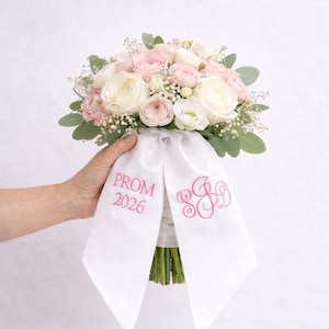 May include: A floral bouquet featuring white and light pink ranunculus flowers, baby's breath, and green eucalyptus leaves. The stems are wrapped in a white satin ribbon with pink embroidery reading PROM 2026 and a decorative monogram design.