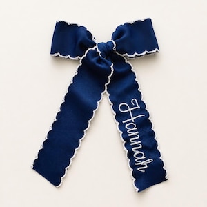 May include: A navy blue hair bow made of ribbed fabric with a white scalloped edge trim. The bow features two long tails, one of which is embroidered with the name Hannah in white cursive script, centered and aligned vertically on the fabric.