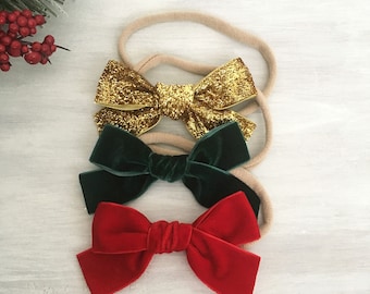 May include: Three hair headbands featuring bows attached to thin, stretchy beige elastic bands. The top bow is gold glitter, the middle bow is dark green velvet, and the bottom bow is bright red velvet, all arranged in a vertical stack.