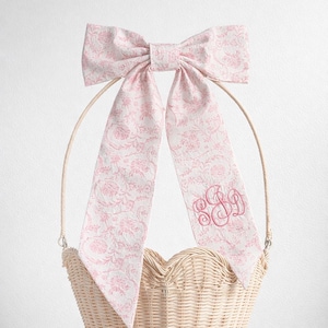 Puede incluir: Una cesta de mimbre con borde en forma de corazón, adornada con un gran lazo rosa con estampado floral. Las largas colas del lazo presentan un monograma rosa. La cesta tiene un asa.