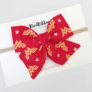 K&ouml;nnte beinhalten: Rote Stoffschleife mit goldenem Wonder Woman Logo und wei&szlig;en Sternen. Befestigt an einem beigefarbenen elastischen Haarband. Auf der wei&szlig;en Karte sind der Text "Kindribbon" und eine Webadresse zu sehen.