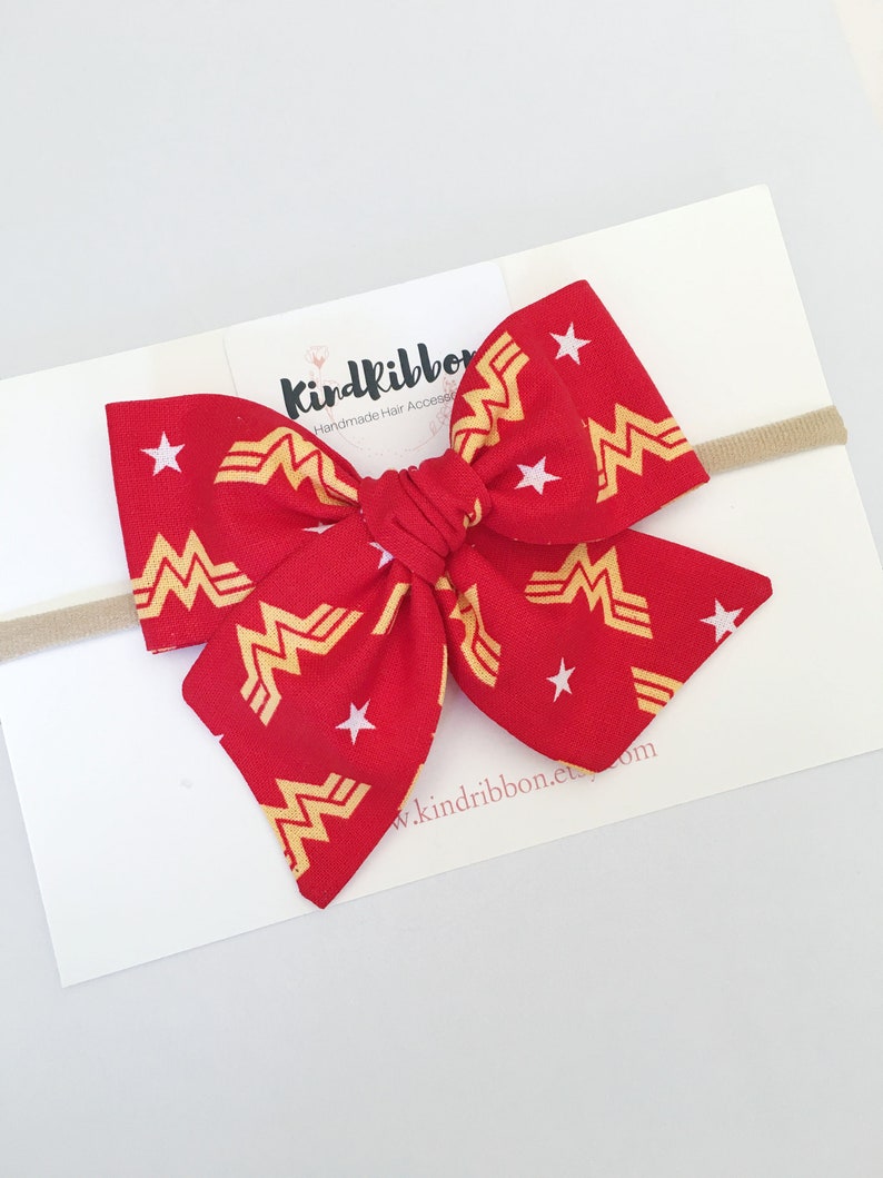 K&ouml;nnte beinhalten: Rote Stoffschleife mit goldenem Wonder Woman Logo und wei&szlig;en Sternen. Befestigt an einem beigefarbenen elastischen Haarband. Auf der wei&szlig;en Karte sind der Text "Kindribbon" und eine Webadresse zu sehen.